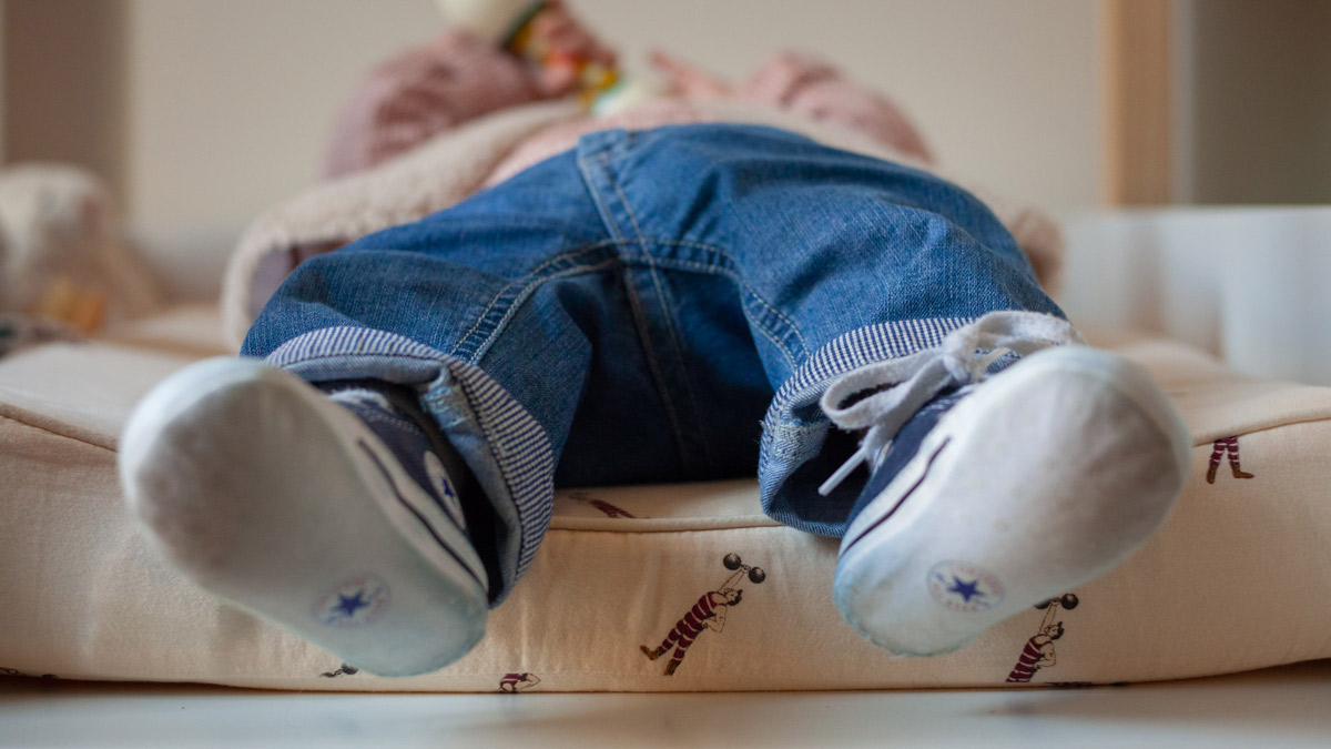 close-up photograph of a baby casually laying down on the changing table, taken at the matress height. He's wearing jeans and a pair of baskets